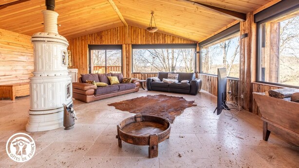 L’intérieur chaleureux d’un chalet avec un poêle à bois, un canapé, un divan en cuir et une grande fenêtre donnant sur le paysage extérieur.