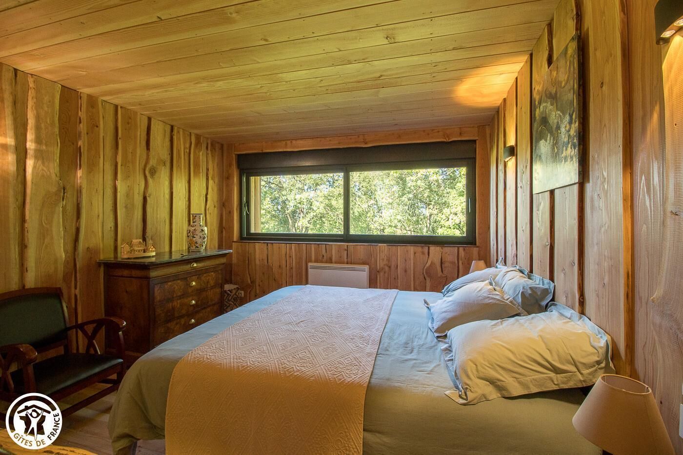 A wooden cabin room with a bed, a dresser, a chair, and a window with a view of trees.