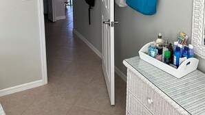 A hallway with a door leading to another room, a coat rack with bags, a wall-mounted shelf with a blue bag, and a wicker basket with toiletries. - Florida Condo for Monthly rental Gated Golf Community available MarchApril 2026. (Fort Myers)