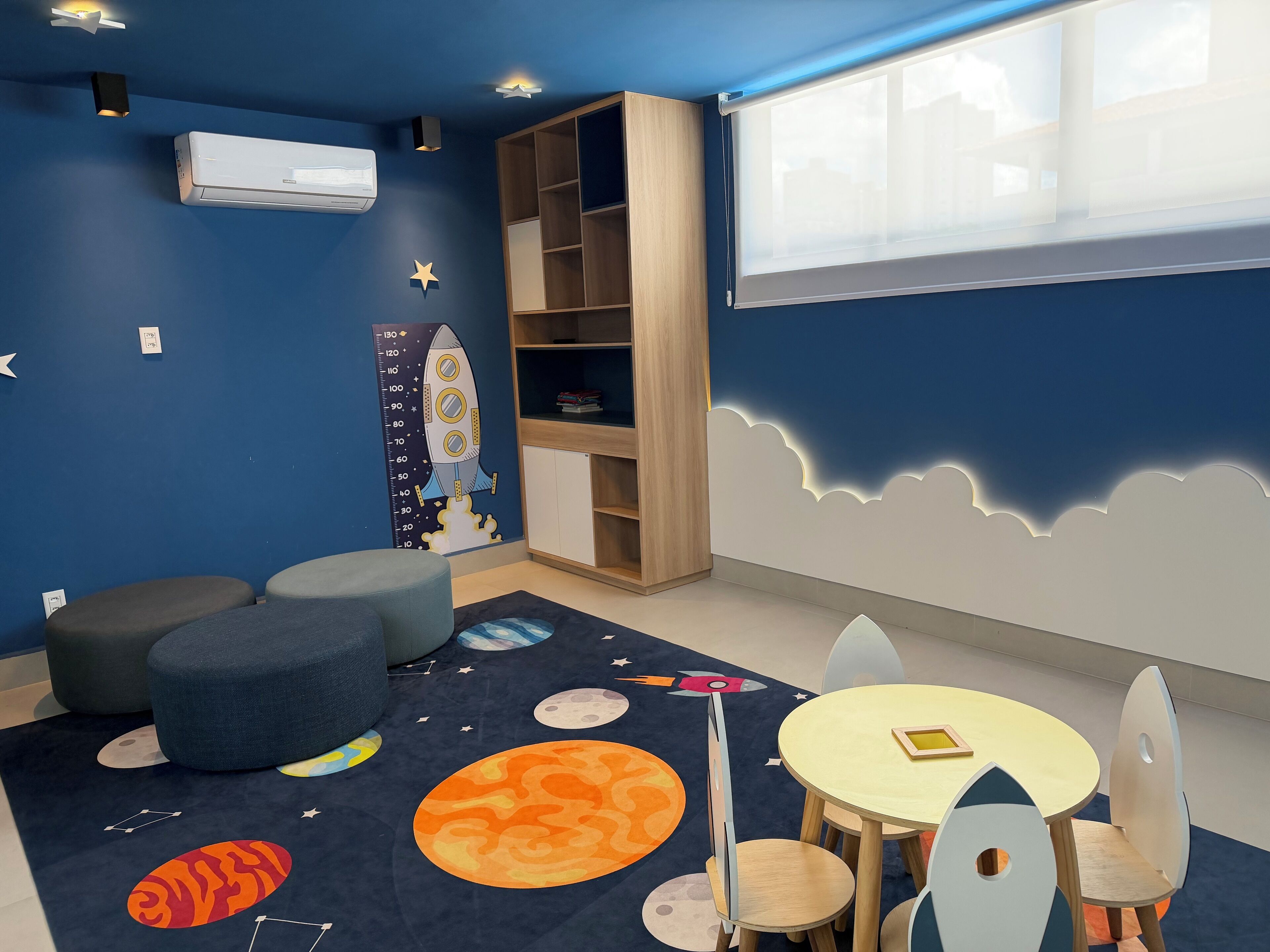 Une pièce avec un mur bleu, un écran de projection, une bibliothèque, une table ronde avec des chaises et un tapis sur le thème de l’espace.