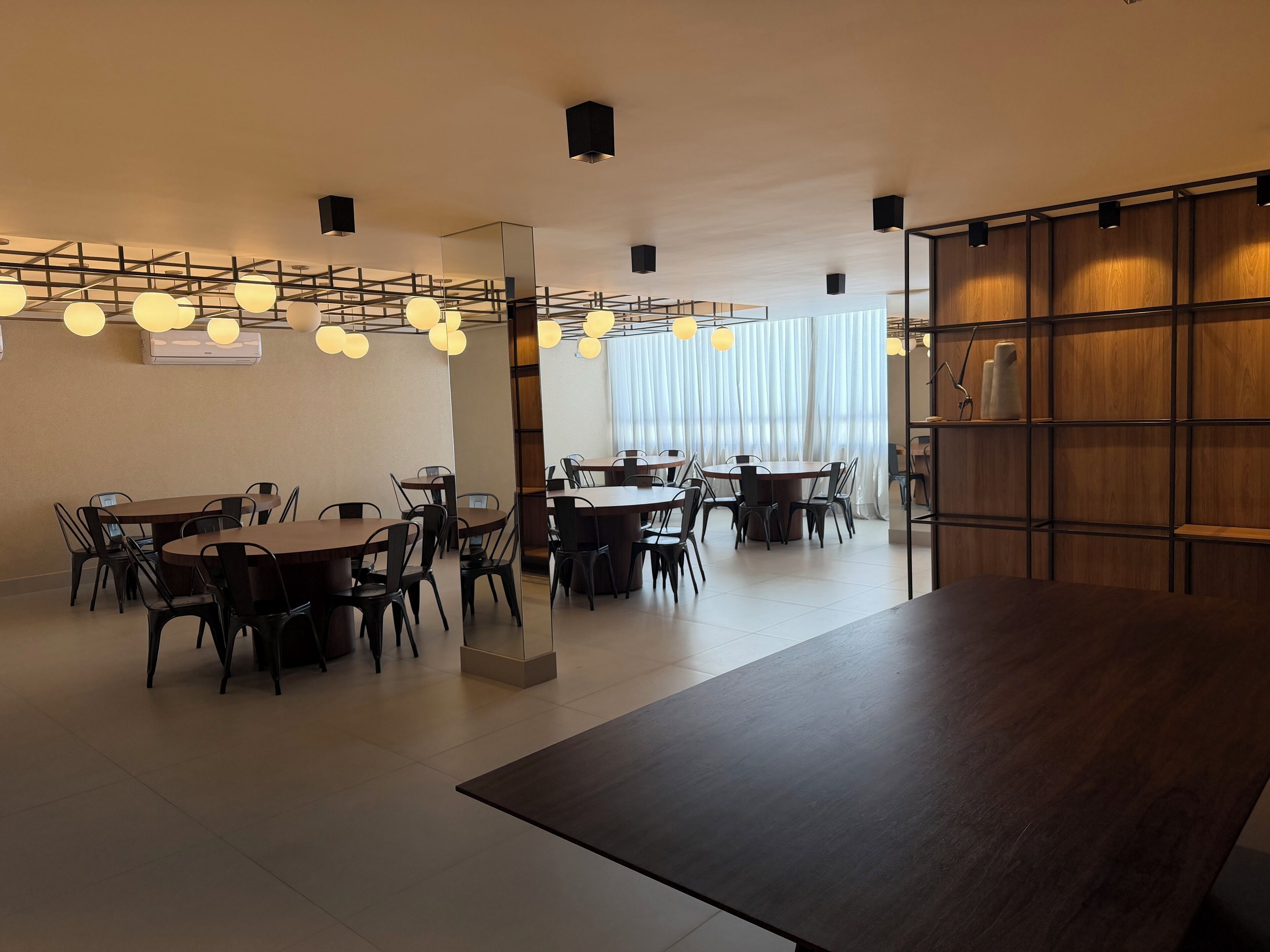 A modern dining area with multiple tables and chairs, a wooden bookshelf, and pendant lighting.