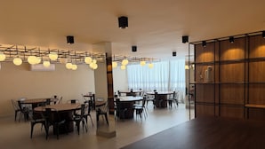 A modern dining area with multiple tables and chairs, a wooden bookshelf, and pendant lighting.
