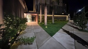 A nighttime view of a building entrance with a well-lit pathway, palm trees, and a grassy area.