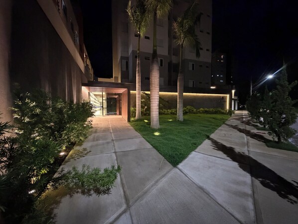 A nighttime view of a building entrance with a well-lit pathway, palm trees, and a grassy area.