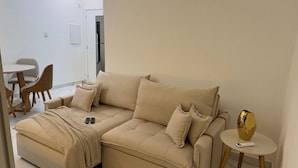 A beige sectional sofa with a blanket and pillows, a small round table with a vase, and a remote control on the sofa.