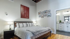 A modern bedroom with a wooden floor, a large bed with a grey and white striped bedspread, a nightstand with a lamp, and a wall-mounted artwork.