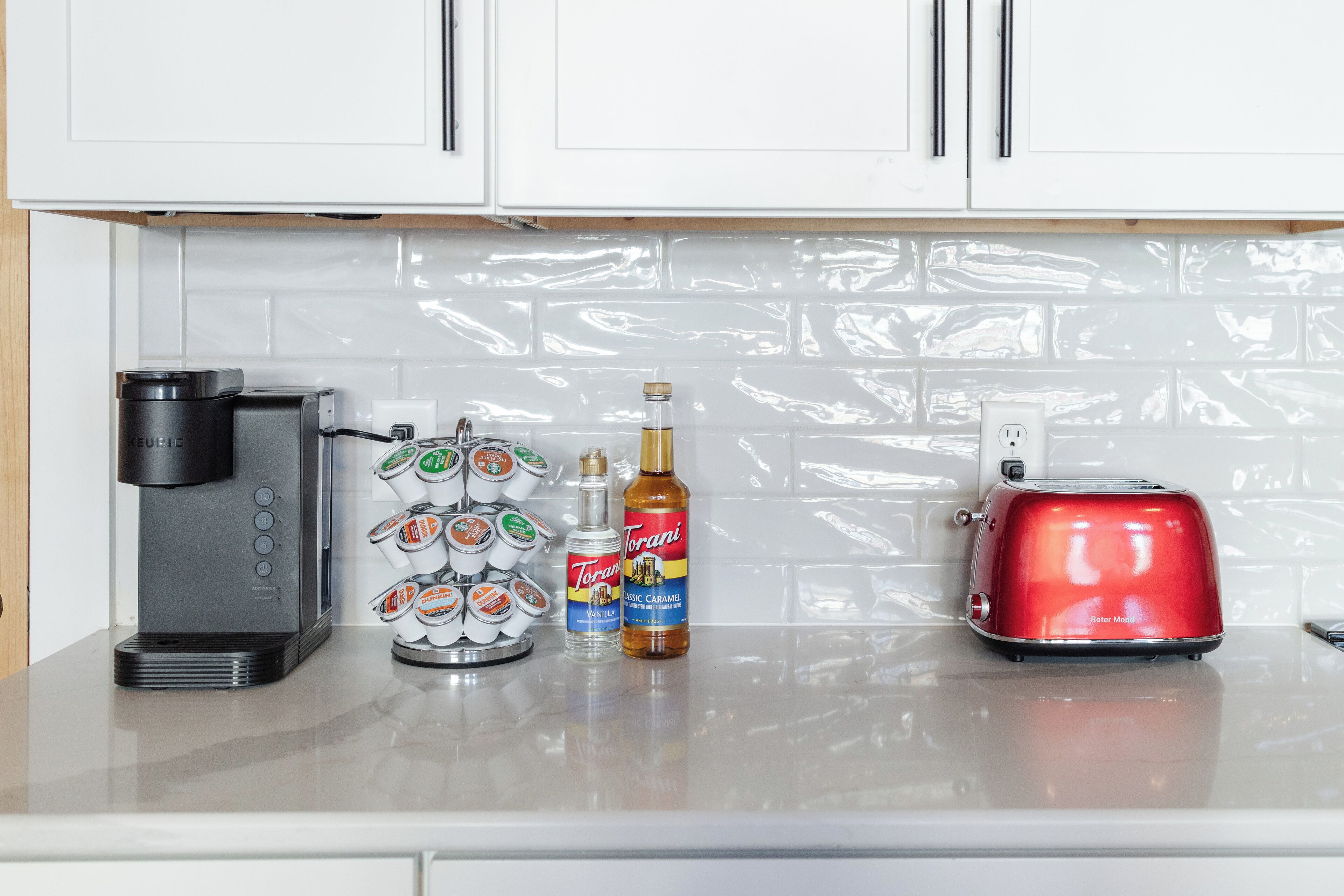 Una encimera de cocina con una cafetera, un dispensador de café, una botella de sirope de caramelo Torani y un tostador rojo.