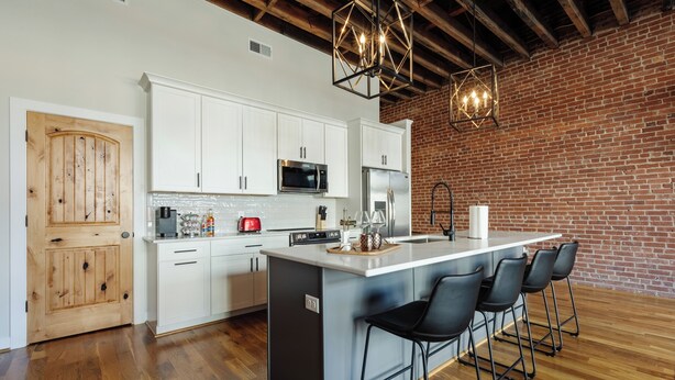 A modern kitchen with white cabinets, a central island with bar stools, a brick wall, and exposed wooden beams.