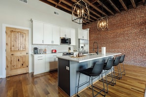 Una cocina moderna con armarios blancos, una isla central con taburetes y una pared de ladrillo, además de vigas de madera a la vista.