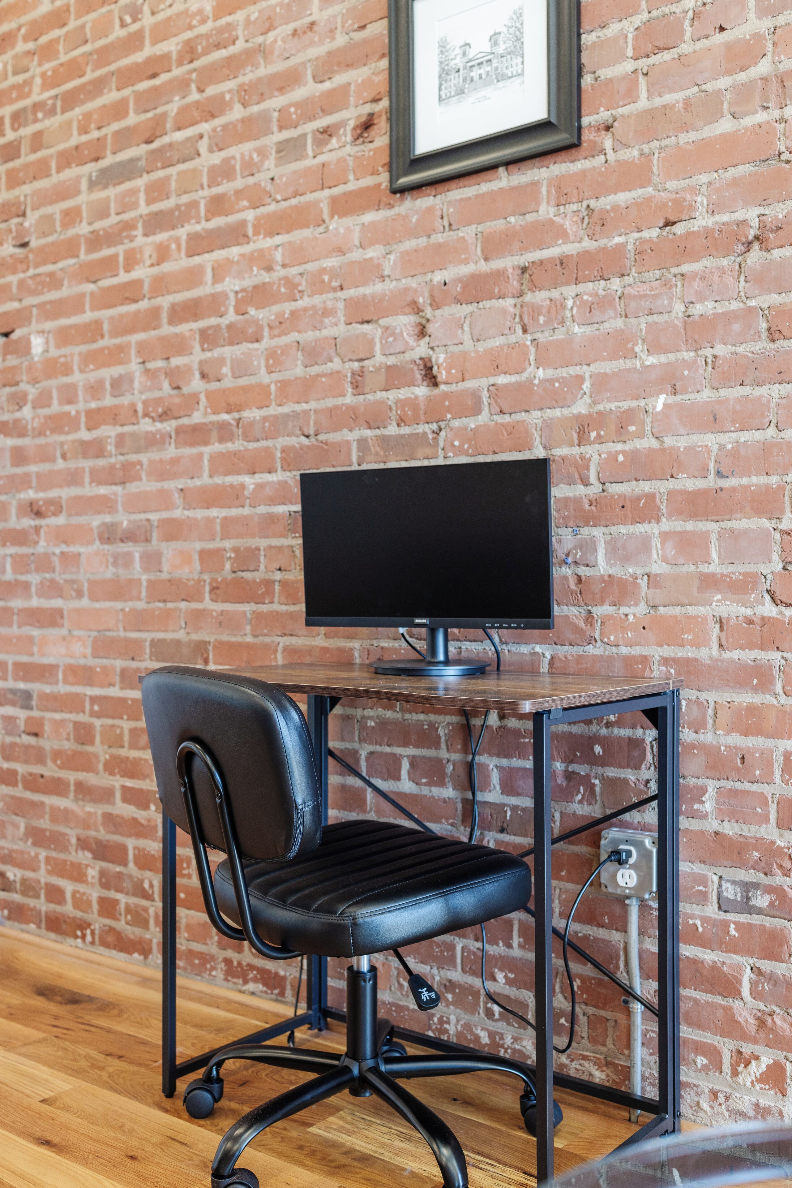 A workspace with a desk, chair, and computer monitor against a brick wall.