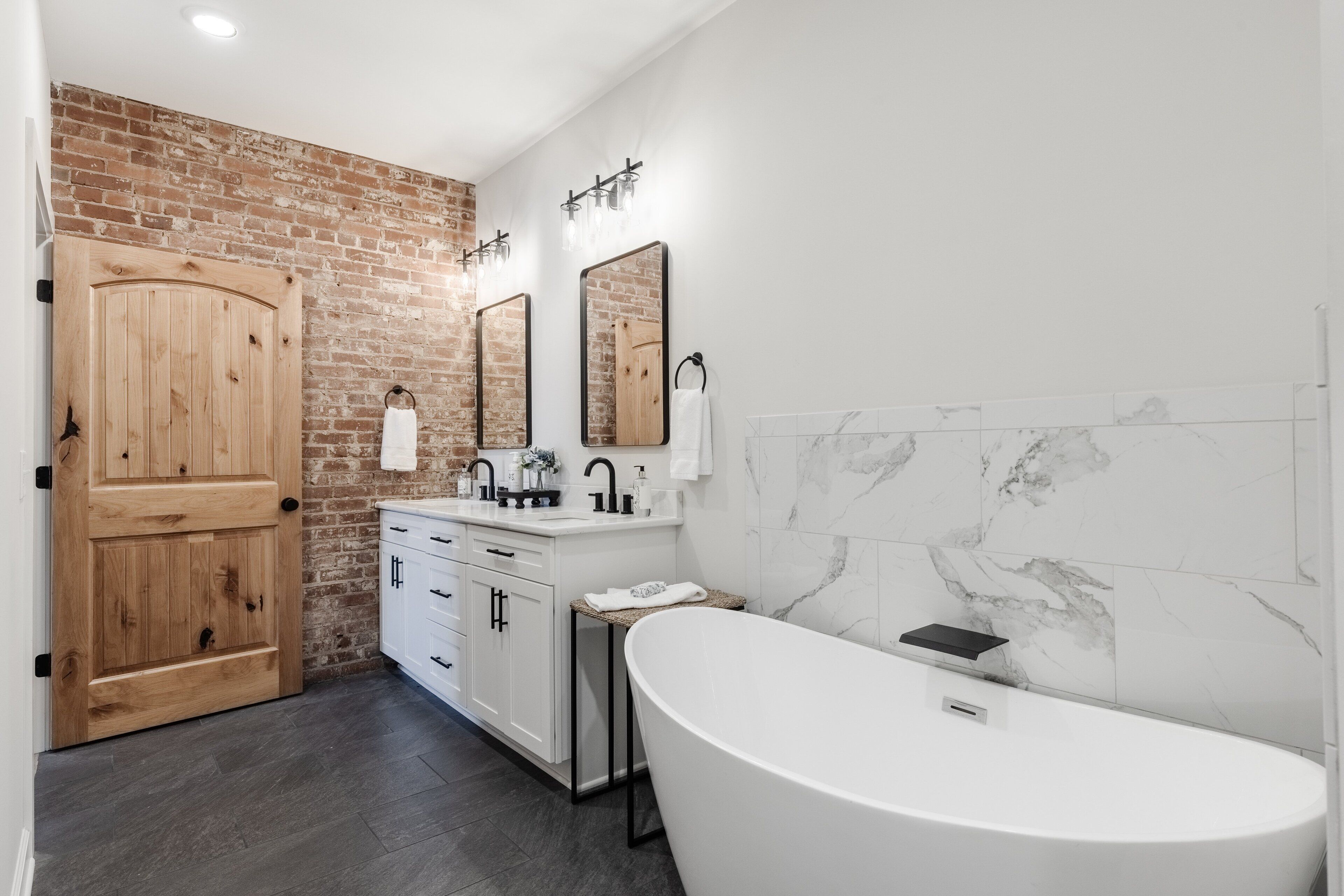 A modern bathroom with a freestanding bathtub, a wooden door, and a brick wall.