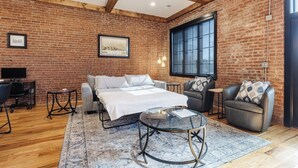 Una habitación con pared de ladrillo, una cama, un sofá, una mesa de centro de vidrio y un escritorio con una computadora.