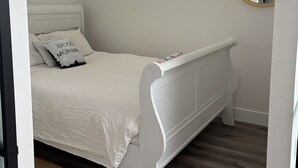 Een slaapkamer met een wit bed, een klein raam, een spiegel en een houten vloer.