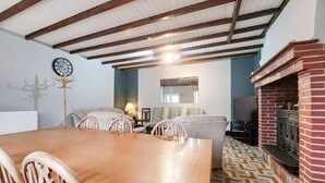 A cozy living room with a wooden dining table, chairs, a sofa, a fireplace, and a ceiling with exposed beams.