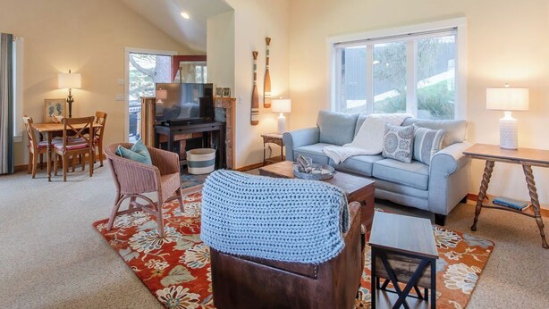 A spacious living room with a sofa, coffee table, and a television. There is a dining area with chairs and a table, and a large window providing natural light. - Haystack Luxury - Private Studio for Monthly Stays, Near Chapman Point, Deck + BBQ (Cannon Beach)