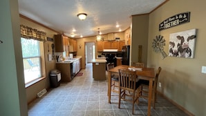 A kitchen with wooden cabinets, a dining table with chairs, a refrigerator, and a window with curtains. - Buck Run Cottage in the heart of Hocking Hills with Wi-Fi & Spectrum TV. (Logan)