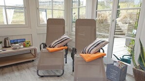 Deux fauteuils inclinables, dotés de coussins rayés et d’oreillers orange, sont placés dans une pièce baignée de lumière, avec de grandes fenêtres donnant sur un jardin.