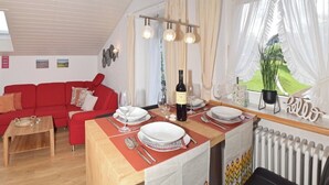 Un salon chaleureux avec un canapé rouge, une table basse en bois et une table à manger dressée avec des assiettes, des verres et des couverts. Une bouteille de vin est posée sur la table, et un radiateur est visible au fond.