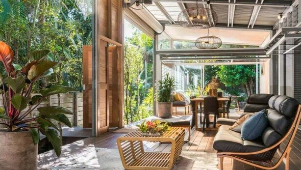 An open-plan living area with a wooden floor, a sofa, a coffee table, and a dining table set with chairs. There are potted plants and a large window providing a view of the outdoors. - Billabelongil - 40A Childe St, Byron Bay (Byron)