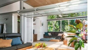 A modern living room with a sofa, a coffee table, and a large window with a view of greenery. - Billabelongil - 40A Childe St, Byron Bay (Byron)
