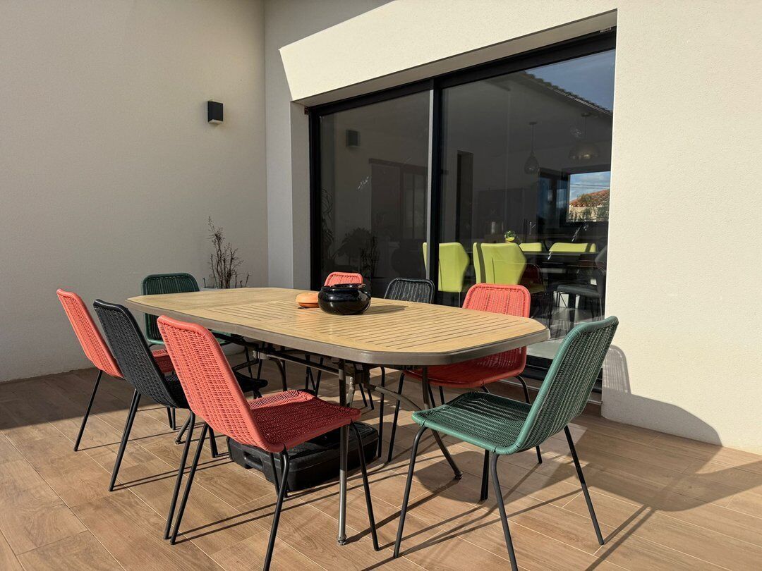 Un espacio de comedor al aire libre con una mesa y sillas de madera, un bol negro y vistas a una casa a través de un vidrio.