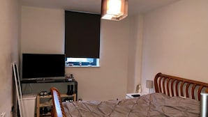 Un dormitorio con cama, televisión, ventana con persianas, escritorio con computadora y una silla.