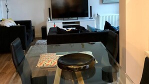 Una sala de estar moderna con una mesa de comedor de vidrio, sillas negras, un televisor de pantalla plana y un mueble blanco para entretenimiento con objetos decorativos.