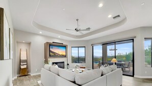 Een moderne woonkamer met een grote hoekbank, een open haard, een flatscreen televisie en uitzicht op een zwembad en bergen door grote glazen schuifdeuren.