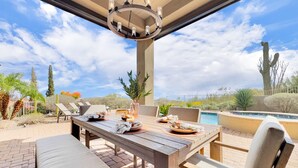 Een terras buiten met een houten eettafel, omringd door stoelen met kussens, een hanglamp en een zwembad op de achtergrond.