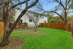 Un jardin à l’arrière d’une maison, avec une terrasse en bois, un petit arbre et une maison blanche.