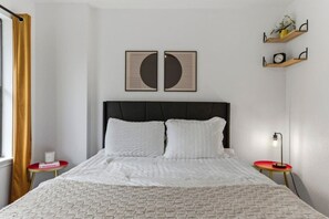 A neatly arranged bedroom with a bed, two bedside tables, a nightstand, a window with curtains, and wall-mounted shelves with decorative items. - Spacious Retreat Near Downtown (Austin)