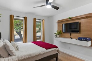 Une chambre à coucher avec un lit, un ventilateur de plafond, un téléviseur fixé sur une étagère en bois et une fenêtre avec des rideaux.