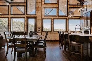 A spacious dining area with a large wooden table and chairs, a kitchen with modern appliances, and large windows offering views of the outdoors. - Heated Outdoor Pool, Video Games, Play-Set, Air Hockey, Rocky Waters (Broken Bow)