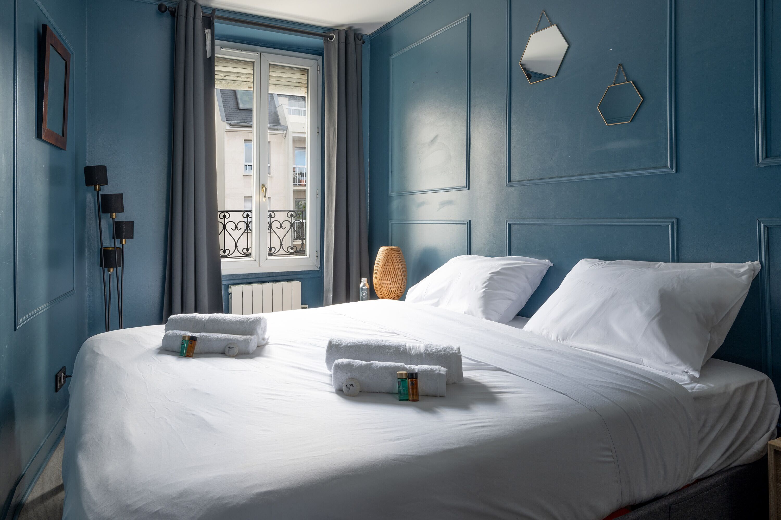Una cama bien hecha con ropa y toallas blancas, una lámpara de noche y una ventana con vista a un balcón.