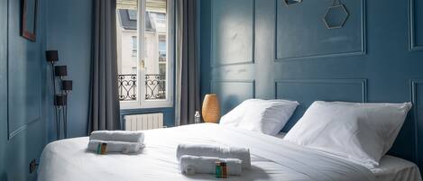 Una cama bien hecha con ropa y toallas blancas, una lámpara de noche y una ventana con vista a un balcón.