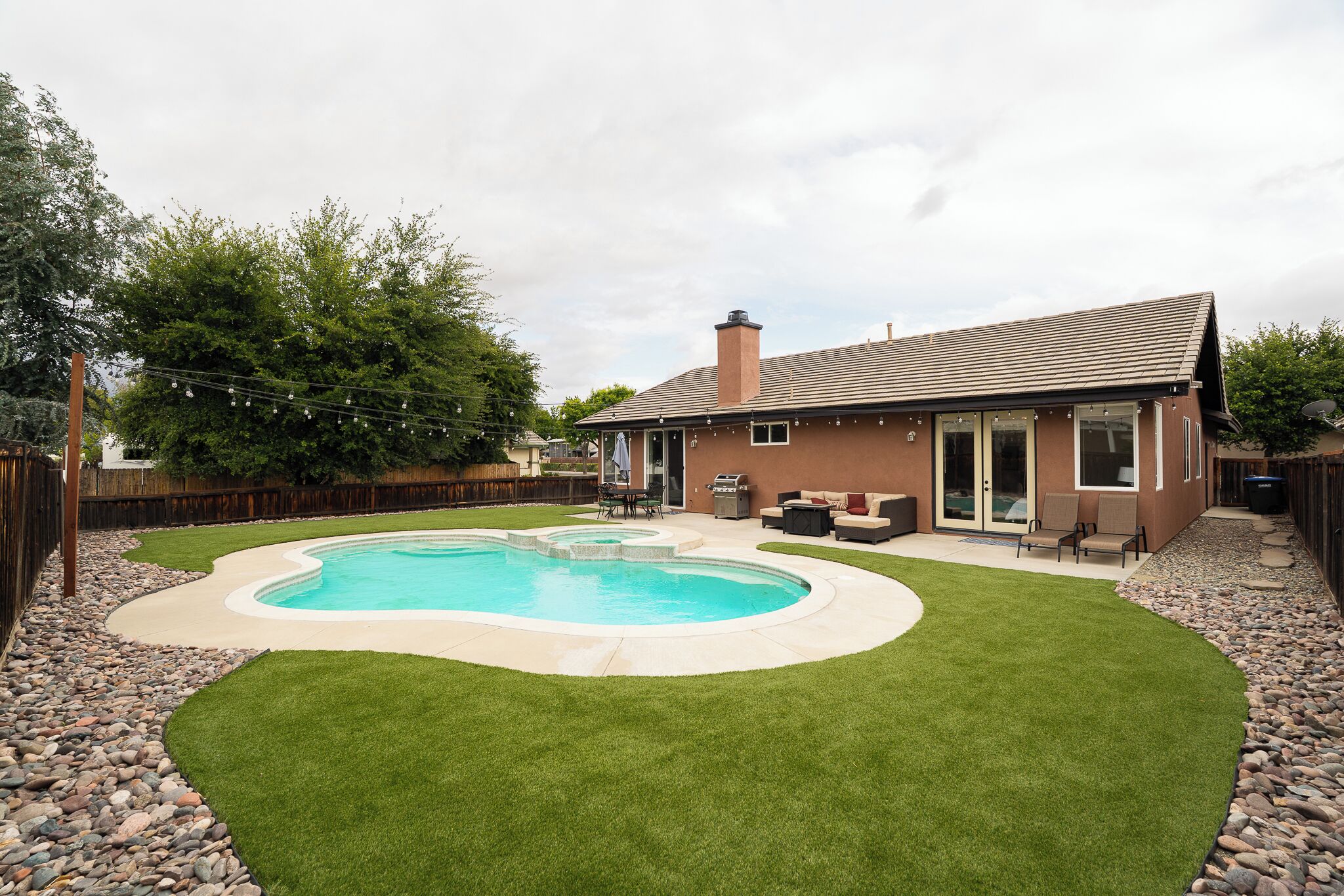 Een achtertuin met een zwembad, een terras met tuinmeubilair, en een huis met een bruin dak en beige gevelbekleding.
