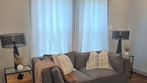 Una sala de estar moderna con un sofá gris, dos mesas auxiliares con lámparas, una mesa de centro y un ventanal con cortinas translúcidas.