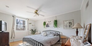 Ein Schlafzimmer mit Bett, Deckenventilator, Fenster mit Vorhängen, Nachttisch, Lampe und Wanddekorationen.