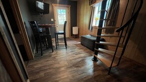 A room with a wooden floor, a dining table with chairs, a sofa, a TV mounted on the wall, and a window with a view of greenery.