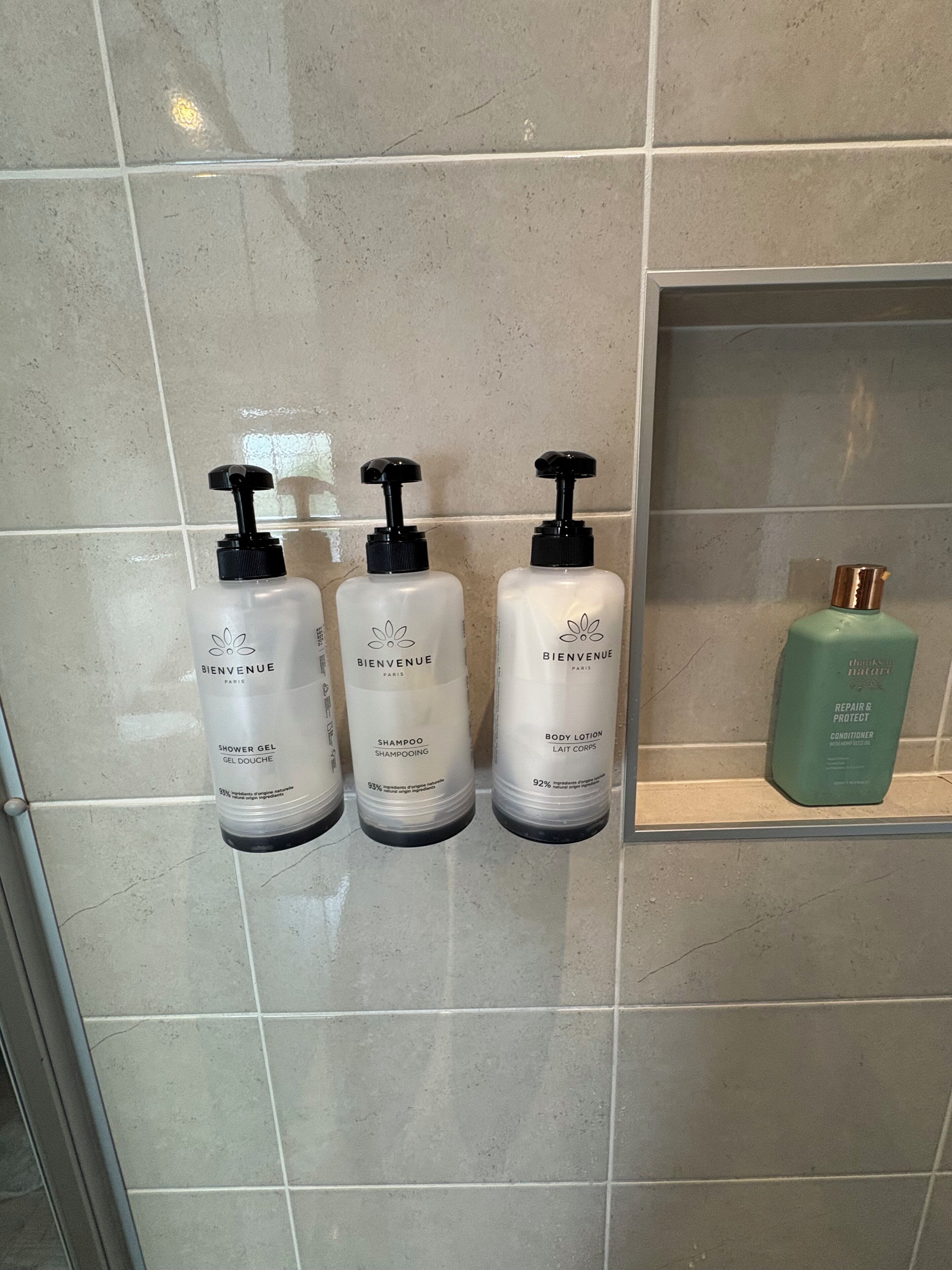 Three bottles of shower gel, shampoo, and body lotion on a tiled wall.