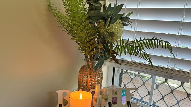A small wooden stool with a lit candle, a potted plant, and a few bottles on a windowsill.