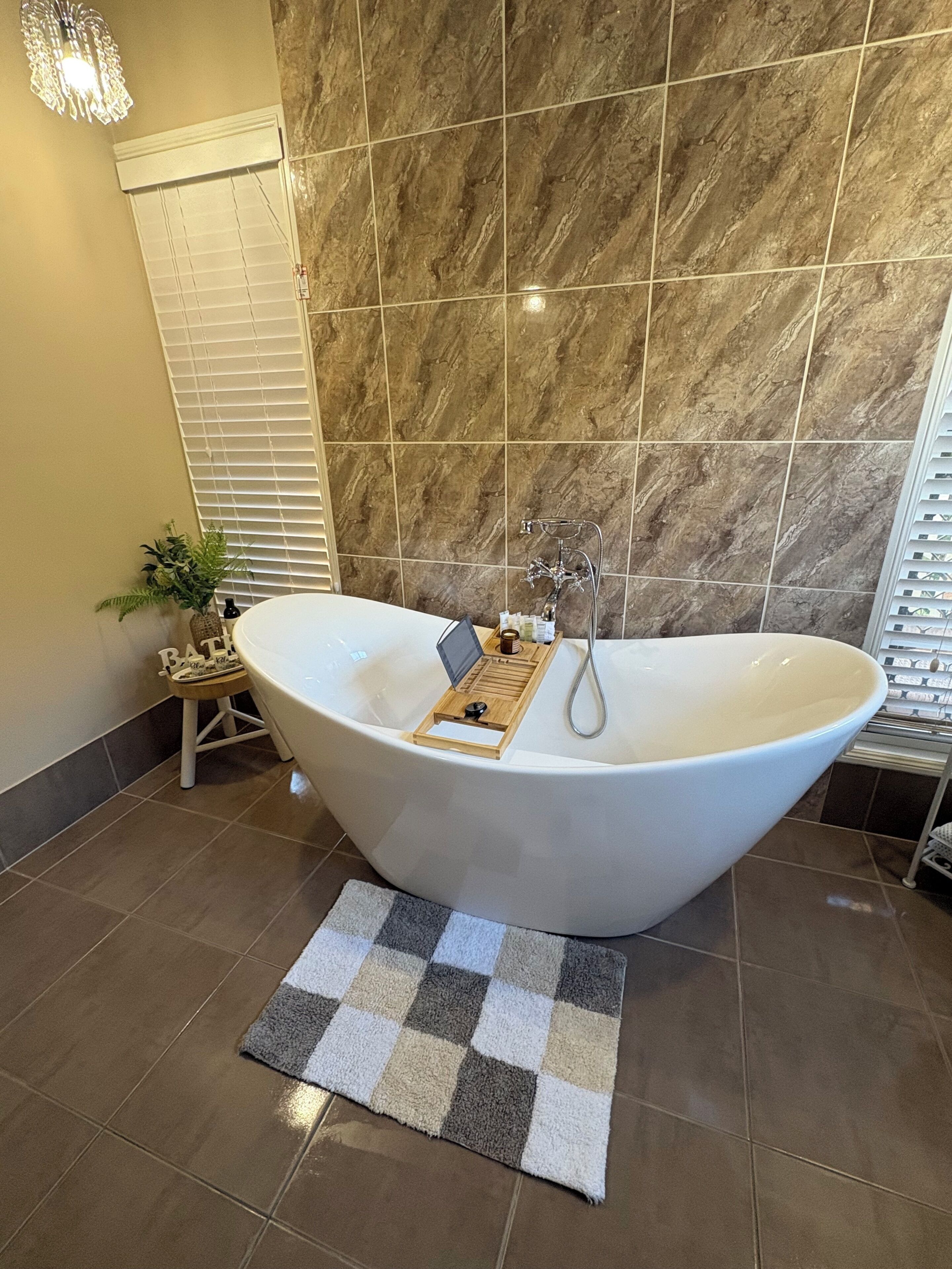 Eine freistehende Badewanne mit Holztablett, einer kleinen Bank und einem karierten Teppich in einem Badezimmer mit Marmorfliesen.