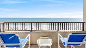 A balcony with two blue lounge chairs and a small table, overlooking a beach with people and a clear sky. - Seawinds 702 (North Myrtle Beach)
