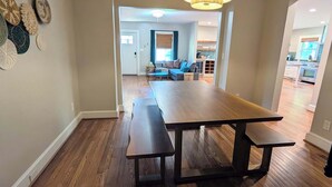 A dining area with a wooden table and benches, a chandelier, and a view into a living room with a sofa and a kitchen in the background. - Stylish Durham Retreat - Walkable, Sleeps 6 (Durham)
