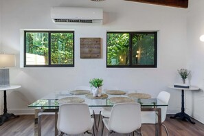 Un coin repas avec une table en verre, des chaises blanches et une vue sur la verdure par les fenêtres.