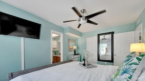 Un dormitorio con cama, ventilador de techo, televisor montado en la pared y un baño visible a través de una puerta abierta.