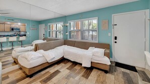 Una sala moderna con un sofá modular, una zona de cocina con microondas y ventanas grandes con persianas.
