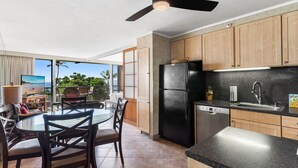 A kitchen with wooden cabinets, a black refrigerator, and a dining area with a glass table and chairs. - Mana Kai 314 (Kihei)