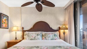A hotel room with a bed, a ceiling fan, a nightstand with a lamp, and a window with curtains. - Mana Kai 314 (Kihei)