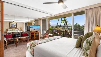 A hotel room with a large bed, a sofa, a TV, and a balcony with a view of palm trees and mountains.